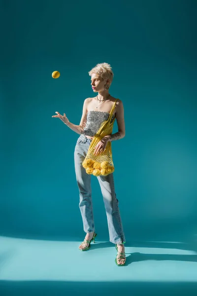 Full length view of tattooed woman in shiny top with sequins holding net bag and throwing in air ripe lemon on blue — Stock Photo