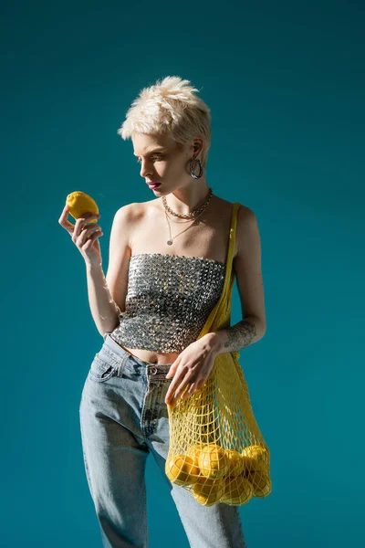 Tattooed albino woman in shiny top with sequins holding net bag with ripe lemons on blue — Stock Photo