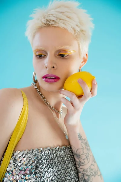 Tattooed woman in shiny top with sequins holding ripe lemon on blue — Stock Photo