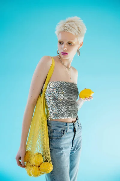 Tattooed woman in shiny top with sequins holding string bag and ripe lemon on blue — Stock Photo