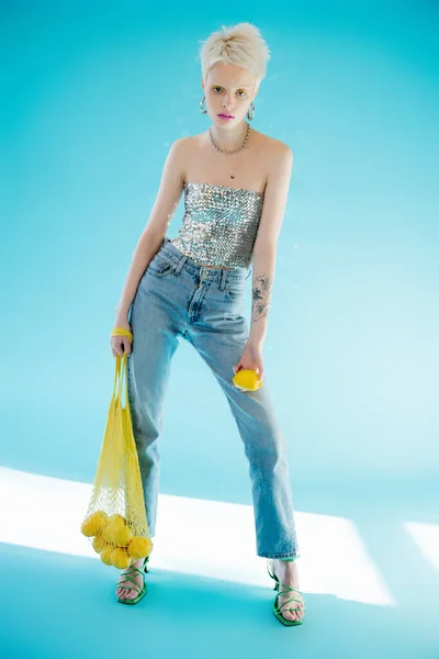 Full length view of tattooed woman in shiny top with sequins holding yellow string bag with ripe lemons on blue — Stock Photo
