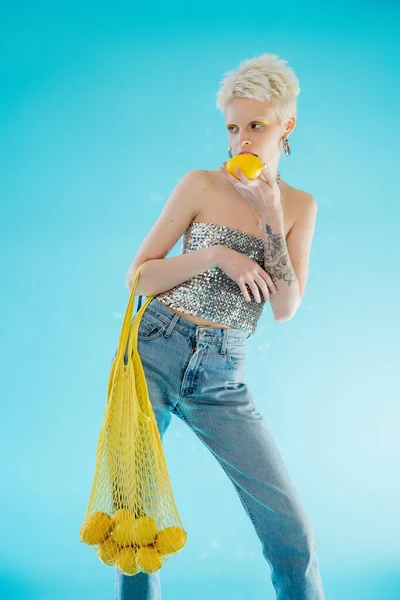 Mulher tatuada em top brilhante com lantejoulas segurando saco de rede com limões orgânicos em azul — Fotografia de Stock