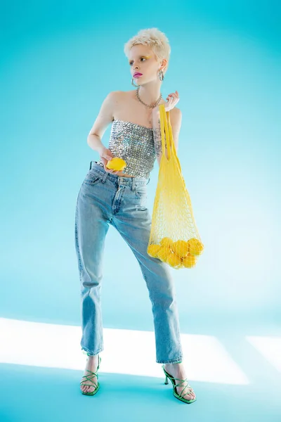 Full length view of tattooed albino woman in shiny top with sequins holding yellow string bag with ripe lemons on blue — Stock Photo