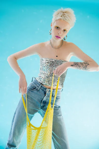 Tattooed woman in shiny top with sequins and denim jeans holding yellow net bag on blue — Stock Photo
