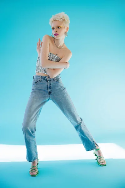 Full length view of tattooed albino model in shiny top with sequins and denim jeans posing on blue — Stock Photo