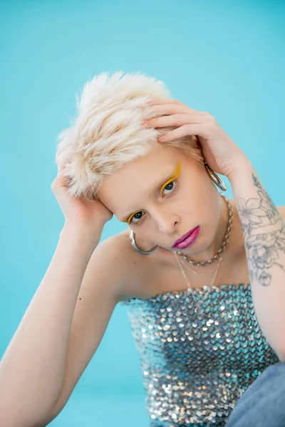Modelo albino tatuado en la parte superior brillante con lentejuelas mirando a la cámara en azul - foto de stock