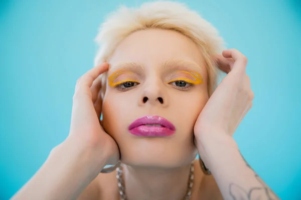Modelo albino rubia con maquillaje brillante mirando a la cámara sobre fondo azul - foto de stock