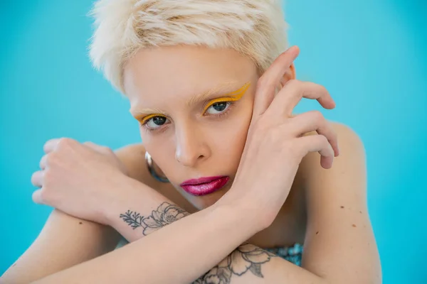Tätowiertes und blondes Albino-Model mit hellem Make-up vor der Kamera auf blauem Hintergrund — Stockfoto