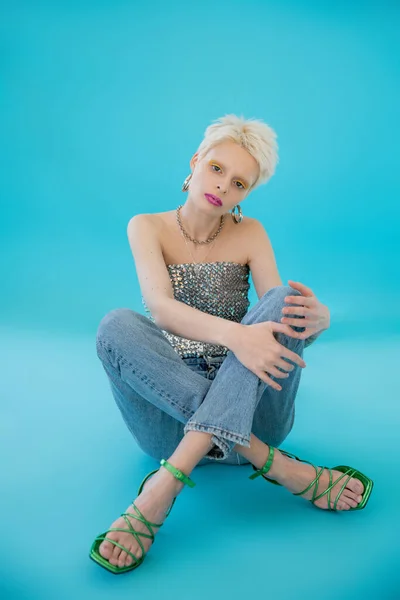 Full length of blonde albino woman with heeled sandals and jeans sitting on blue — Stock Photo