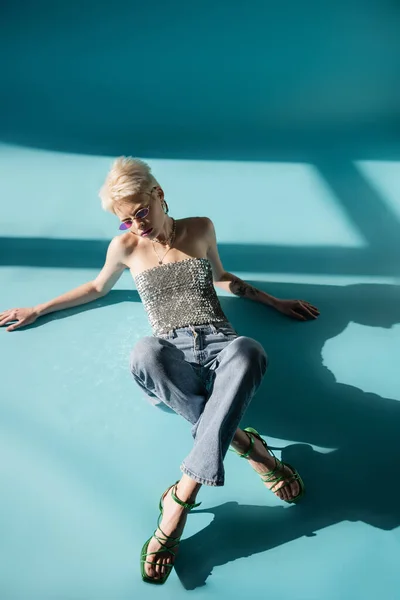 Ansicht einer tätowierten Albino-Frau mit hochhackigen Sandalen und Jeans, die auf blau posiert — Stockfoto