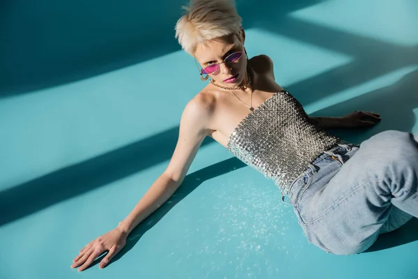 Vista aérea del modelo albino tatuado en gafas de sol rosadas mirando a la cámara en azul - foto de stock