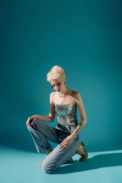 Longitud completa de modelo albino tatuado en gafas de sol de moda y traje posando sobre fondo azul - foto de stock