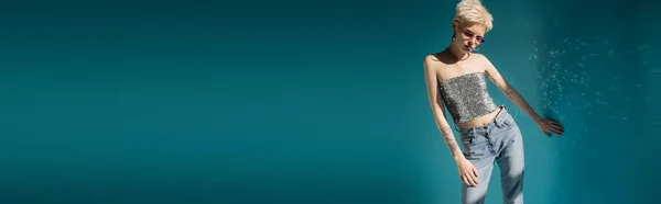 Татуйована модель альбіно в модних сонцезахисних окулярах і стильному вбранні позує біля синьої стіни, банер — стокове фото
