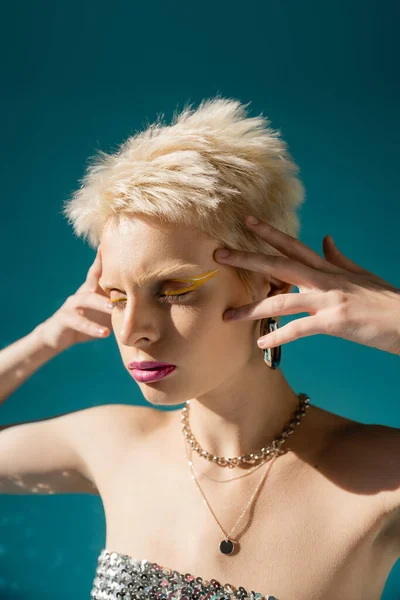 Portrait of blonde albino woman with trendy makeup posing with hands near head on blue — Stock Photo