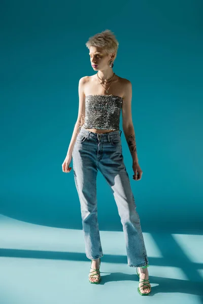 Full length view of tattooed albino model in top with sequins and denim jeans standing on blue — Stock Photo