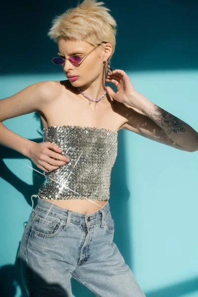 Luz solar no corpo do modelo albino tatuado em óculos de sol rosa e top brilhante com lantejoulas no fundo azul — Fotografia de Stock