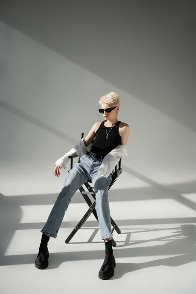 Volle Länge der stilvollen blonden Frau in Sonnenbrille und trendigem Outfit sitzt auf Klappstuhl auf grau — Stock Photo