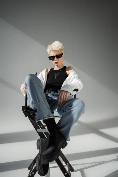 Полная длина стильной блондинки в модных солнцезащитных очках и черных сапогах, сидящих на раскладном стуле на сером — стоковое фото