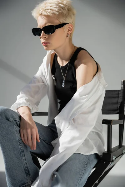 Donna bionda con occhiali da sole alla moda e vestiti alla moda seduta sulla sedia pieghevole su grigio — Foto stock