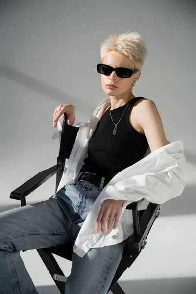 Modelo rubia en gafas de sol de moda y ropa elegante sentado en silla plegable en gris - foto de stock
