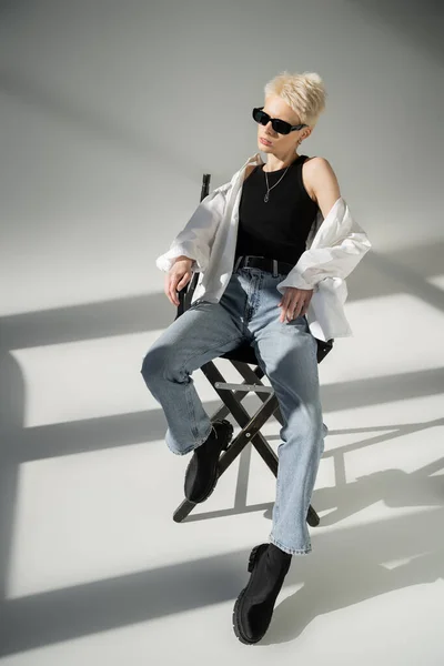 Pleine longueur de femme blonde élégante en lunettes de soleil noires assis sur chaise pliante sur gris — Photo de stock
