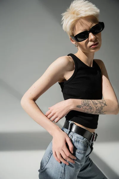 Stylish blonde woman with tattoo posing in black sunglasses on grey — Stock Photo