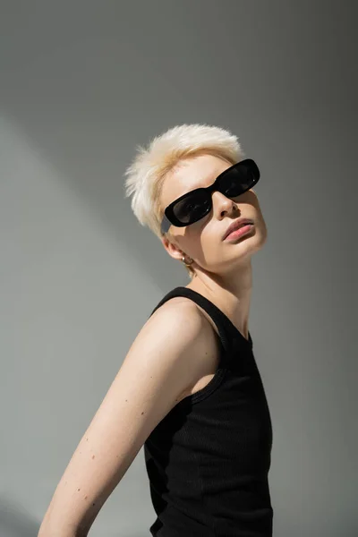 Стильная блондинка в майке и черные солнцезащитные очки на сером — стоковое фото