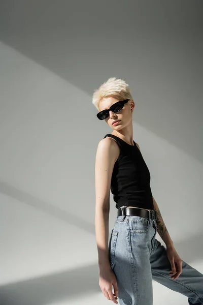 Стильная блондинка в майке и джинсах в черных солнцезащитных очках на сером фоне — стоковое фото