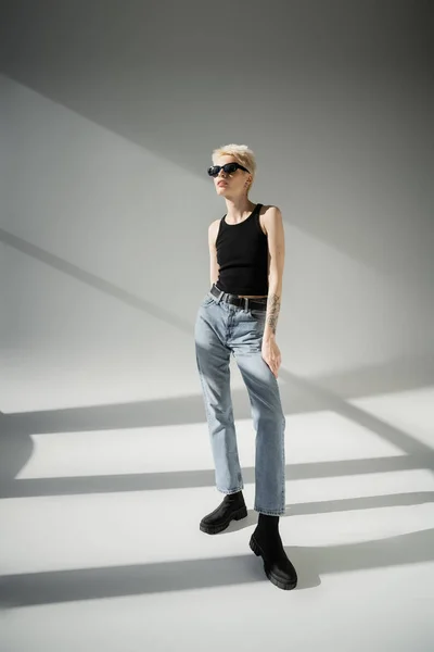 Повна довжина татуйованої моделі в чорних сонцезахисних окулярах і джинсах, позує на сірому фоні — стокове фото