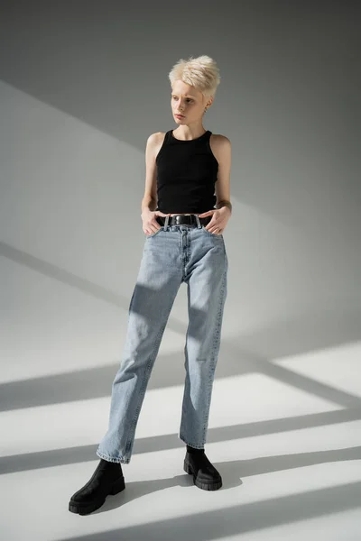 Longitud completa de mujer albina en camiseta negra y jeans posando con las manos en bolsillos sobre fondo gris - foto de stock