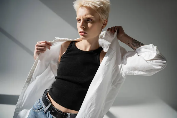 Pretty blonde woman with tattoo on hand wearing white shirt on grey background with shadows — Stock Photo