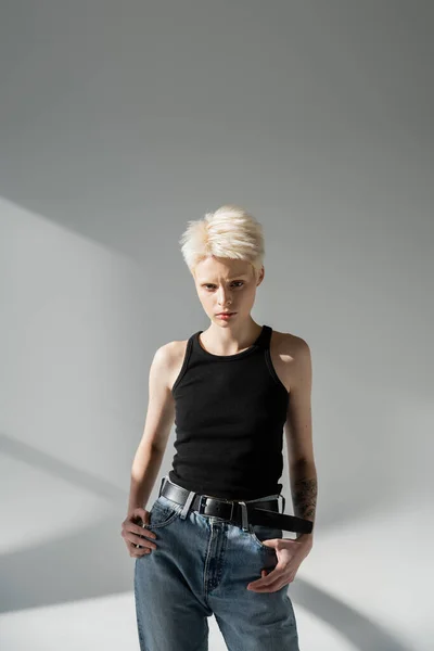Ernsthafte Albino-Frau mit Tätowierung auf der Hand posiert in Jeans und Tank-Top auf grau — Stockfoto