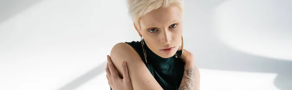 Молодая альбиноса с татуировкой на руке обнимает себя на сером фоне с тенями, баннером — стоковое фото