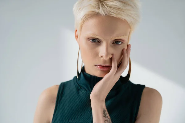 Mujer albina tatuada con la cara de piel clara tocando con la mano mientras mira la cámara en gris - foto de stock