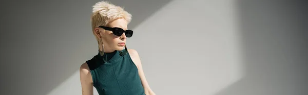 Blonde woman in sunglasses posing while standing in green dress on grey background, banner — Stock Photo