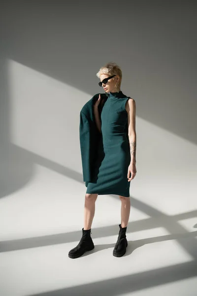 Volle Länge der tätowierten und blonden Frau mit Sonnenbrille posiert in grünem Kleid und Mantel auf grauem Hintergrund — Stockfoto