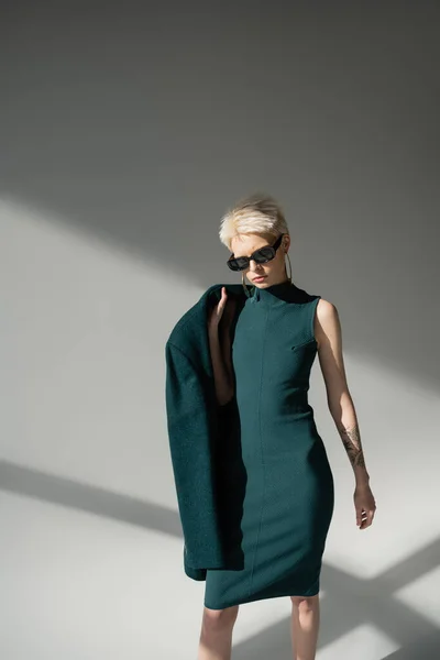 Блондинка в стильных солнцезащитных очках позирует с зеленым пальто на плече на сером фоне — стоковое фото
