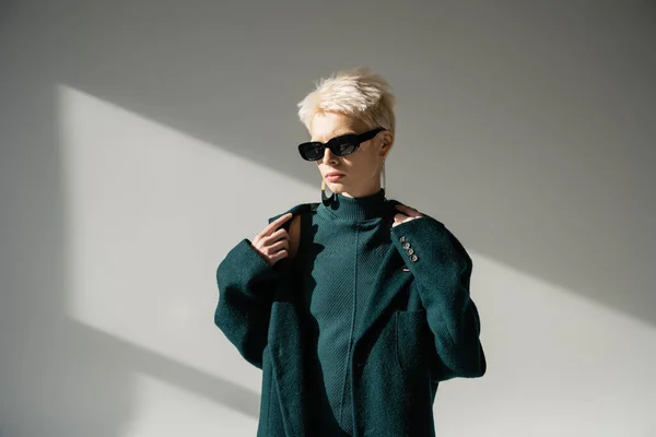 Блондинка в модних сонцезахисних окулярах в зеленому пальто на сірому фоні — стокове фото