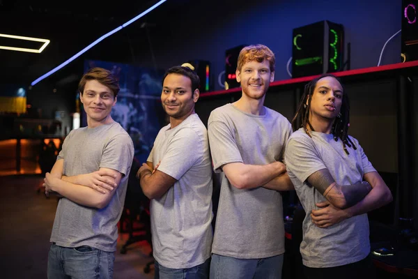 Smiling multiethnic gamer team in grey t-shirts crossing arms in gaming club — Stock Photo