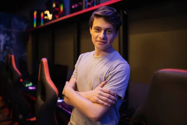 Улыбающийся молодой геймер, скрещивающий руки и смотрящий в камеру в размытом игровом клубе — стоковое фото