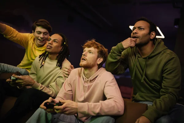 QUIIV, UCRÂNIA - FEVEREIRO 13, 2023: Homem indiano focado sentado perto de amigos inter-raciais alegres jogando videogame no clube de jogos — Fotografia de Stock