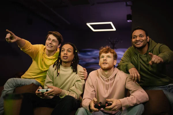 KYIV, UKRAINE - FEBRUARY 13, 2023: Smiling interracial men pointing with fingers while friends playing video game in gaming club — Stock Photo