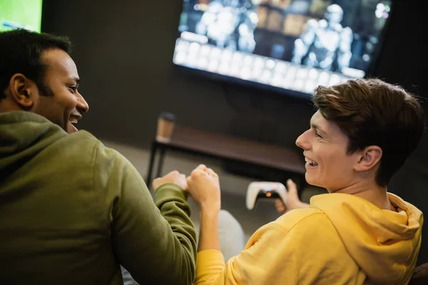 KYIV, UKRAINE - FEBRUARY 13, 2023:Side view of cheerful multiethnic friends doing fist bump while playing video game in gaming club — Stock Photo