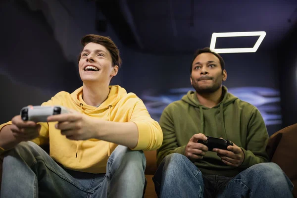 KYIV, UKRAINE - FEBRUARY 13, 2023: Low angle view of cheerful man playing video game with indian friend in gaming club — Stock Photo