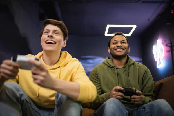 KYIV, UKRAINE - FEBRUARY 13, 2023: Low angle view of positive indian man playing video game with blurred friend in gaming club — стокове фото