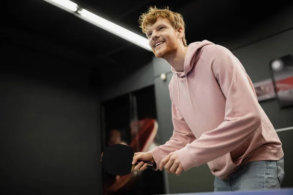 Низкий угол обзора улыбающегося рыжего человека, играющего в настольный теннис в игорном клубе — стоковое фото