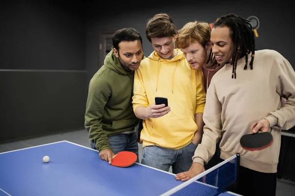 Smiling interracial friends using smartphone near table tennis in gaming club — Stock Photo