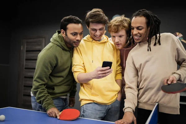 Amigos multiétnicos sonriendo mientras usan el teléfono inteligente cerca del tenis de mesa en el club de juegos - foto de stock