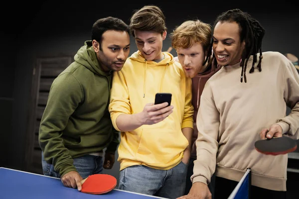 Alegre amigos multiétnicos utilizando el teléfono celular cerca de tenis de mesa en el club de juegos - foto de stock