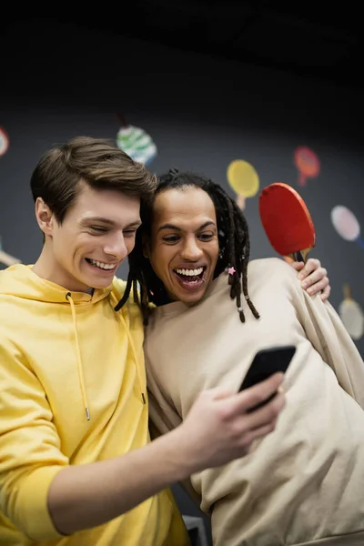 Hombre sonriente usando un teléfono inteligente y sosteniendo raqueta de tenis cerca de amigo multirracial en el club de juegos - foto de stock